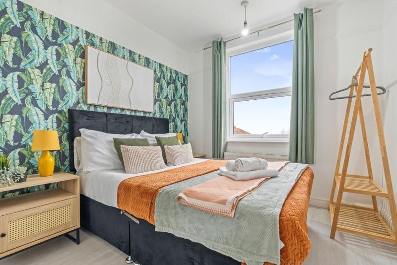 Bright bedroom with double bed, tropical wallpaper headboard, wooden furniture and green curtains