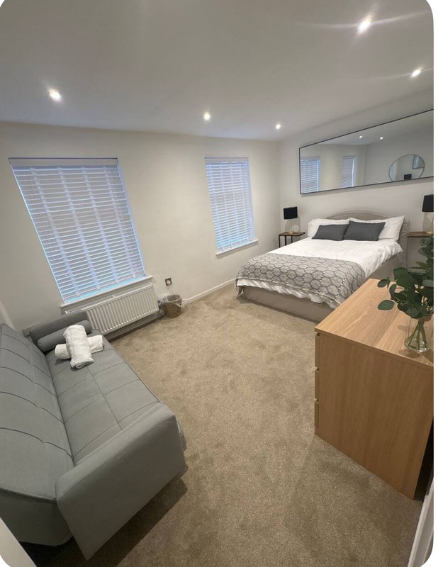 Modern bedroom with double bed, grey sofa, wooden dresser, and large windows with blinds