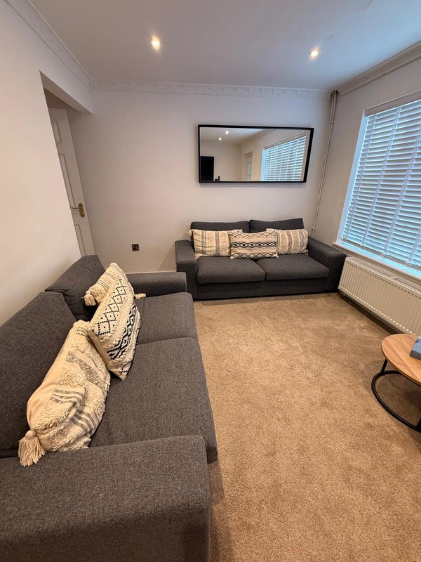 Living room with two dark grey sofas, patterned cushions, wall-mounted mirror, and large window with blinds