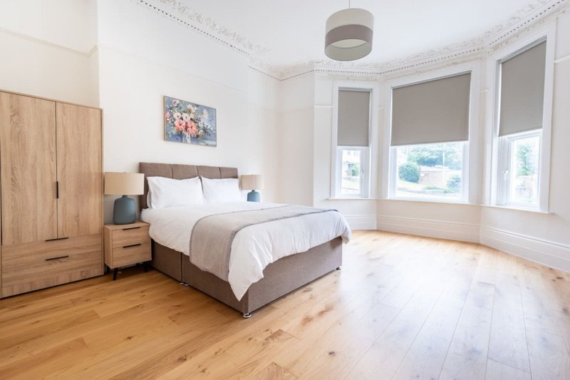 Bright bedroom with double bed, wooden wardrobe, and large bay windows with roller blinds
