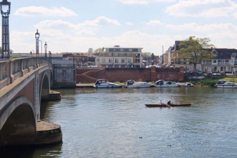 Riverside view with stone bridge, historic buildings, parked cars and water in foreground