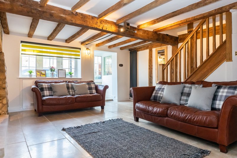 Open-plan living room with wooden beams, two burgundy leather sofas, grey rug and wooden staircase