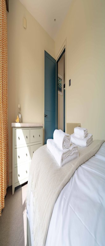 Narrow bedroom with single bed, white bedding, towels, small cabinet and open door