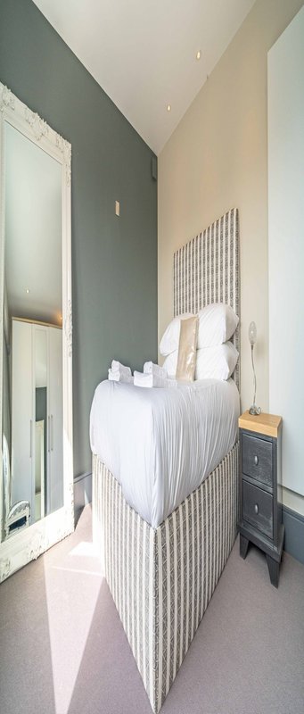 Narrow bedroom with white crib, grey walls, mirror, and wooden nightstand
