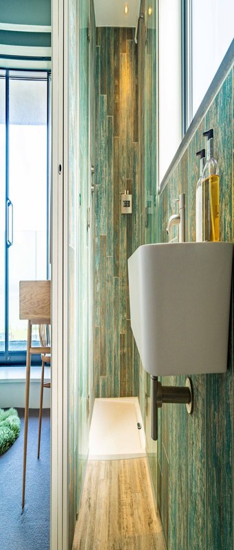 Narrow hallway with green-tiled walls, wall-mounted sink, and view through to wooden flooring and seating area