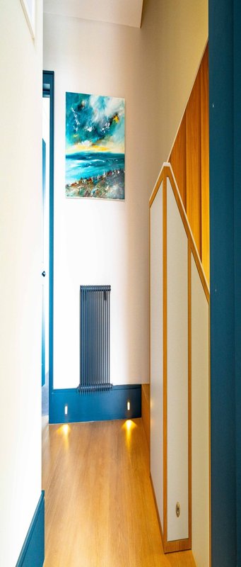 Narrow hallway with wooden floor, blue wall panels, framed coastal artwork, and wooden door frame