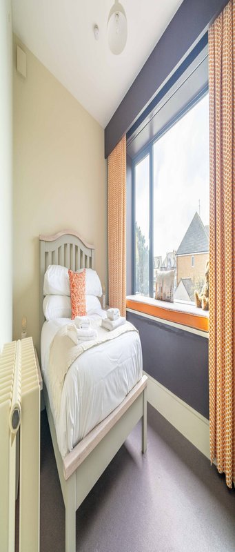 Narrow bedroom with single bed, large window with orange curtains, white walls and wooden headboard