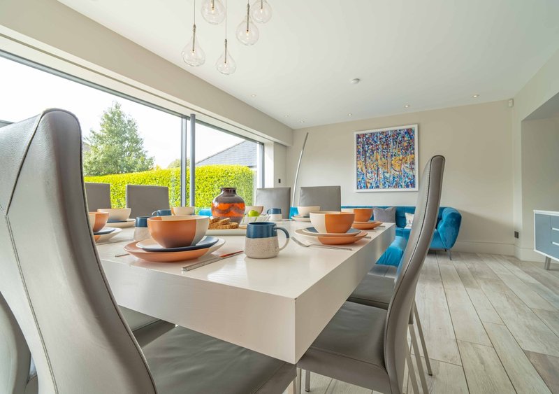Modern open-plan dining area with white table, grey chairs, orange tableware, and large windows overlooking garden