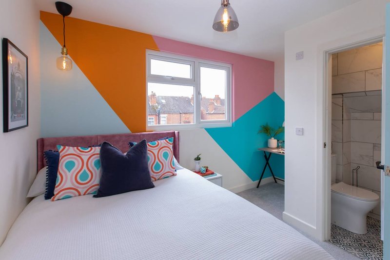 Modern bedroom with colorful geometric wall art, double bed, patterned cushions, pendant lighting, and ensuite bathroom visible