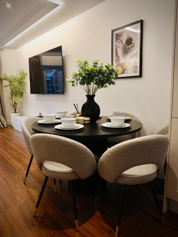 Dining area with black round table, two beige upholstered chairs, place settings and flower arrangement