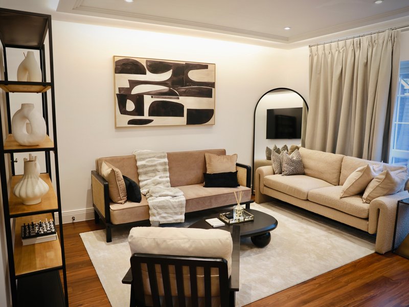 Modern living room with tan sofas, abstract wall art, wooden shelving, and large windows with cream curtains