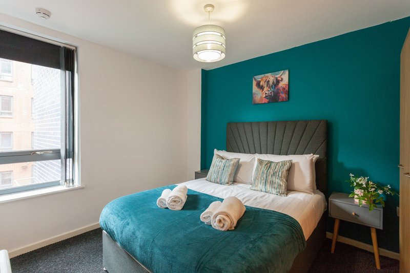 Modern bedroom with teal accent wall, double bed with rolled towels, and window with city view