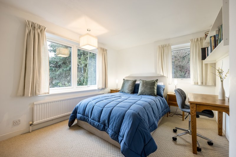 Bright bedroom with blue duvet, wooden desk, radiator and large windows with cream curtains