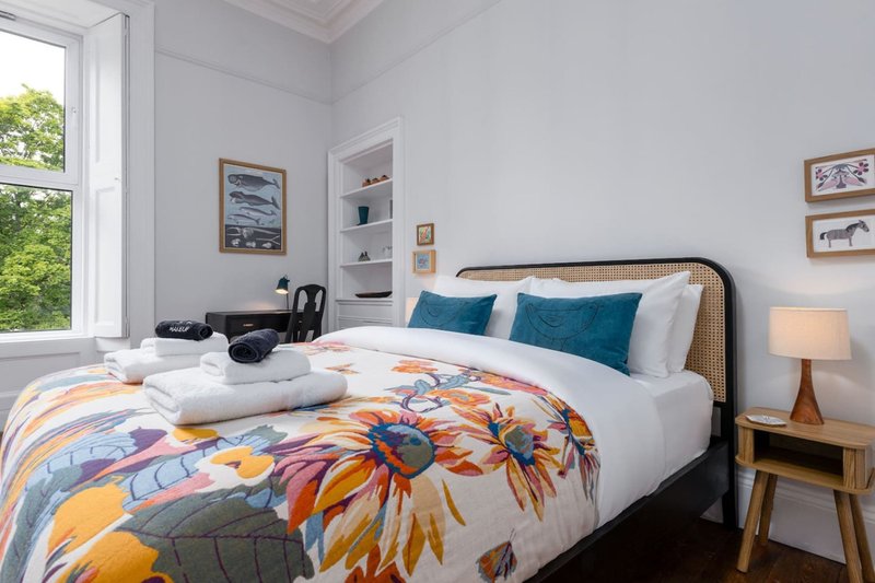 Bright bedroom with double bed, colorful floral duvet, rattan headboard, and window overlooking garden