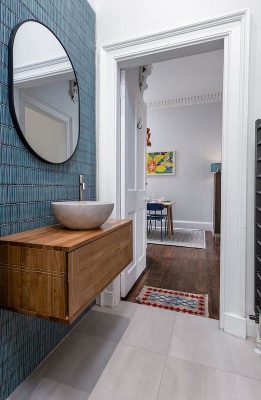 Modern bathroom with blue tile wall, wooden vanity, round mirror, and view into hallway