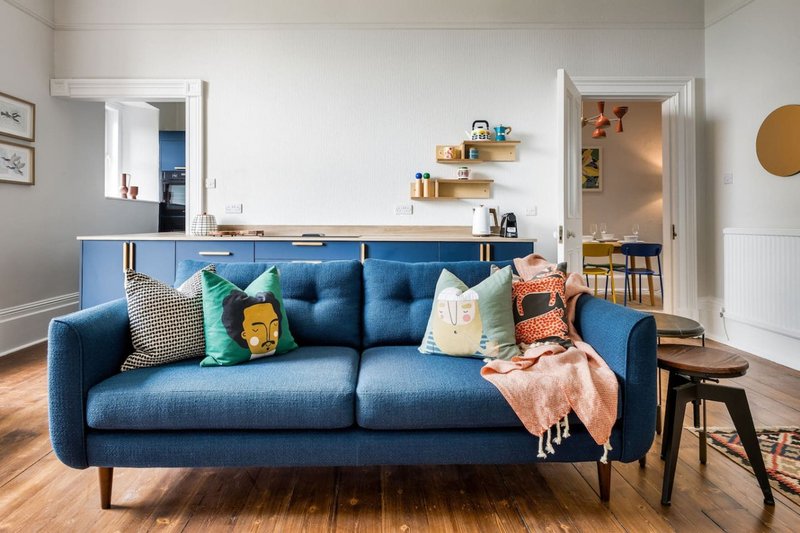 Modern living room with blue sofa, colorful cushions, wooden floor and open-plan kitchen