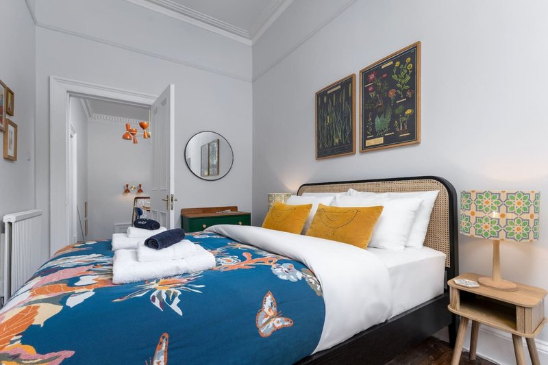 Bedroom with double bed, blue floral duvet, yellow pillows, wooden nightstand and wall art