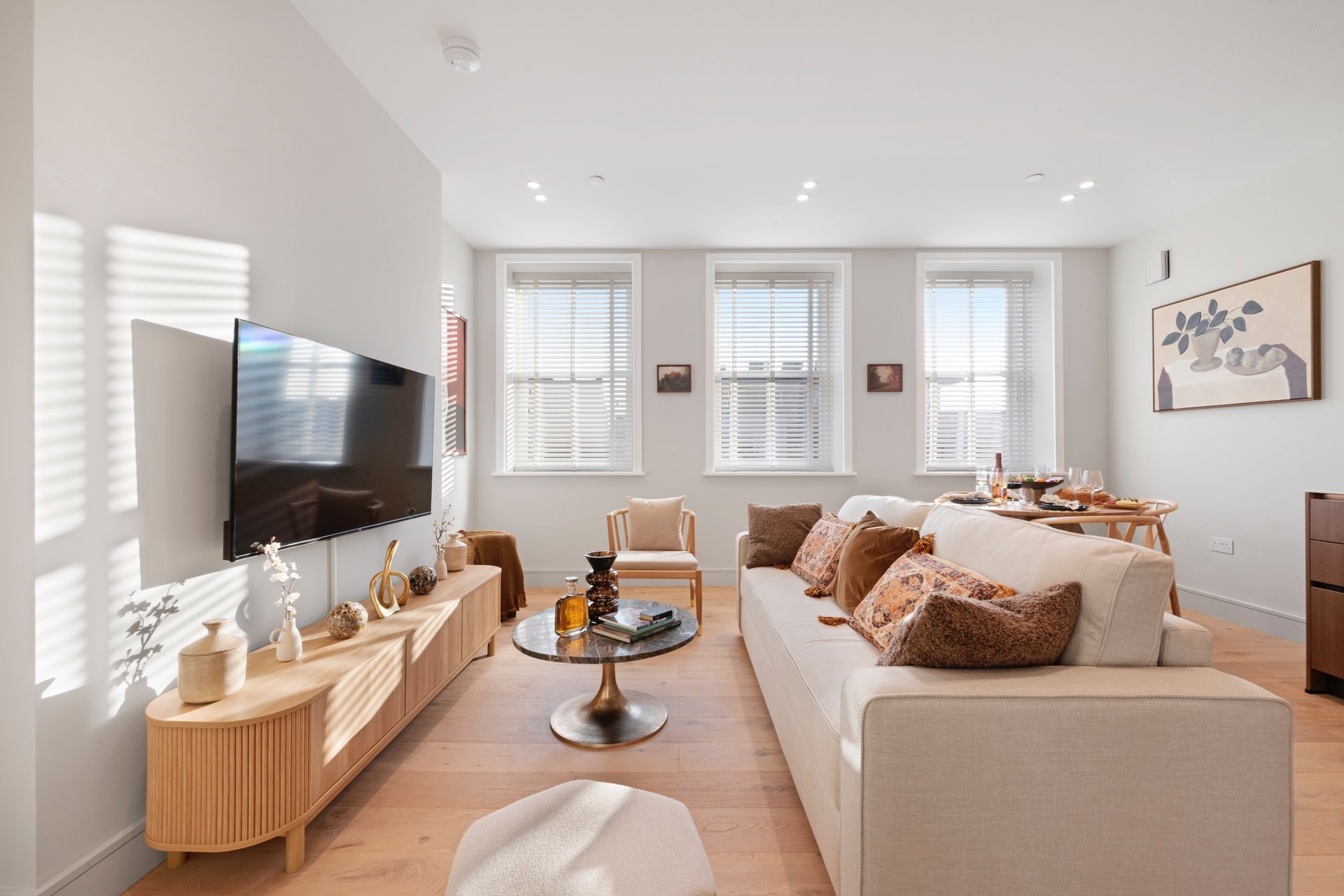Bright living room with cream sofa, wooden TV stand, and three large windows with natural light