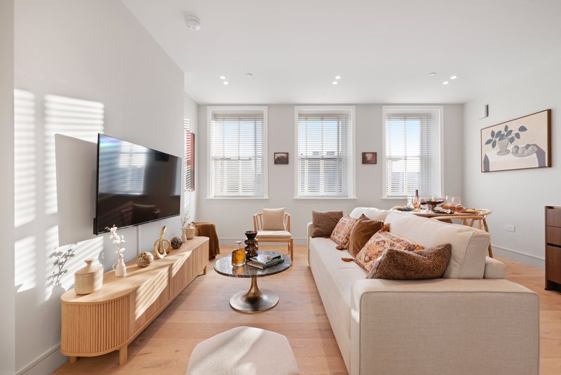 Bright living room with cream sofa, wooden TV stand, and three large windows with natural light