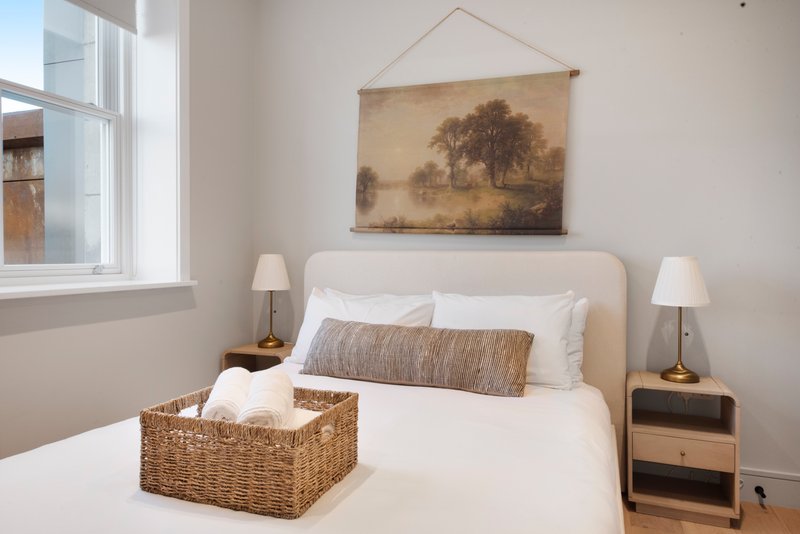 Bright bedroom with double bed, woven storage basket, bedside tables and landscape artwork