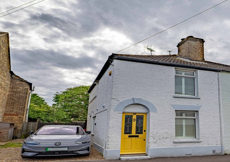 White cottage with yellow door, driveway with parked car, brick chimney visible