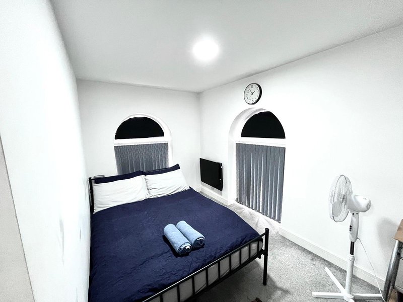 Bedroom with single bed, dark blue bedding, white pillows, wall clock and standing fan