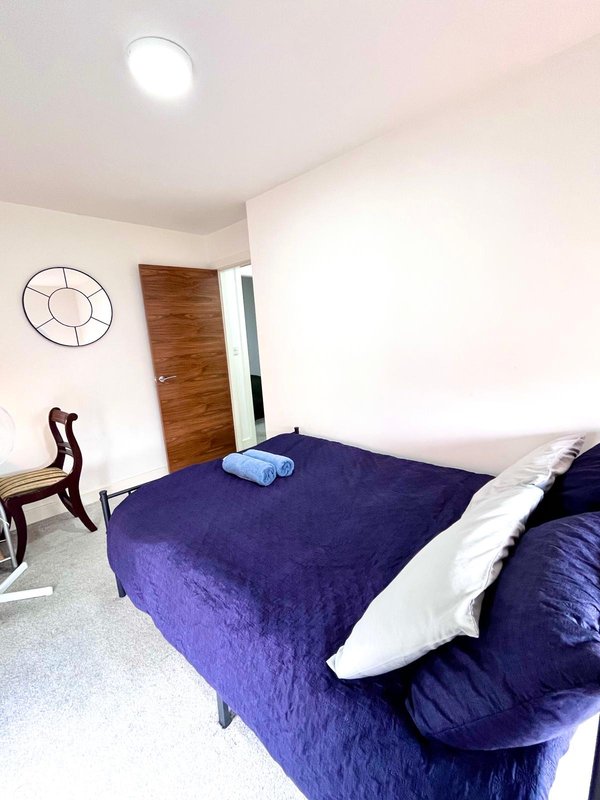 Bedroom with double bed, purple bedding, wooden door, wall clock and recessed ceiling light