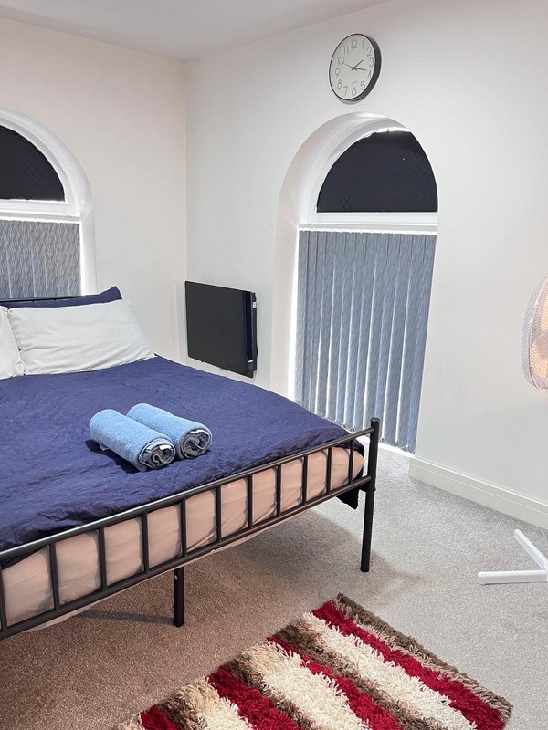 Bedroom with single metal-frame bed, blue bedding, rolled towels, wall clock, and striped rug
