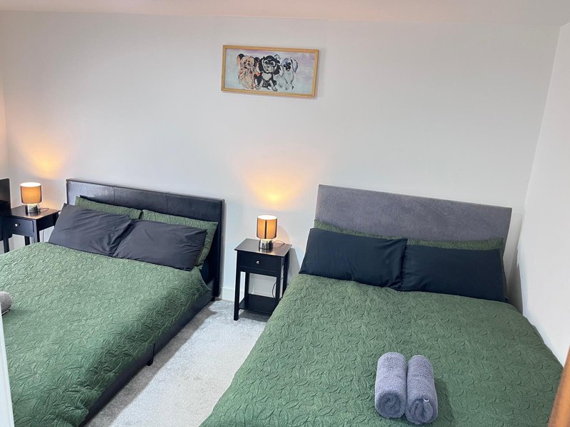 Twin bedroom with green quilted bedding, dark headboards, bedside tables with lamps, and rolled towels