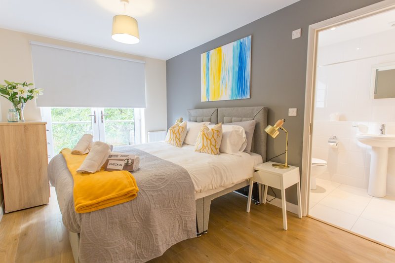 Modern bedroom with grey upholstered bed, yellow accents, ensuite bathroom visible, wooden flooring
