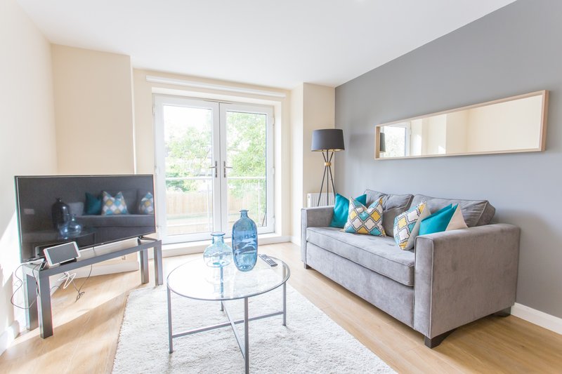 Bright living room with grey sofa, TV on stand, round glass table and patio doors