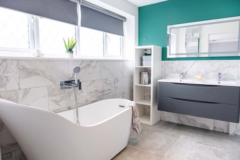 Modern bathroom with freestanding white bathtub, marble tile walls, teal accent wall and vanity sink.
