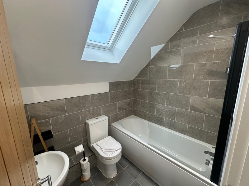 Modern bathroom with grey tile walls, white toilet, bathtub with shower screen and skylight