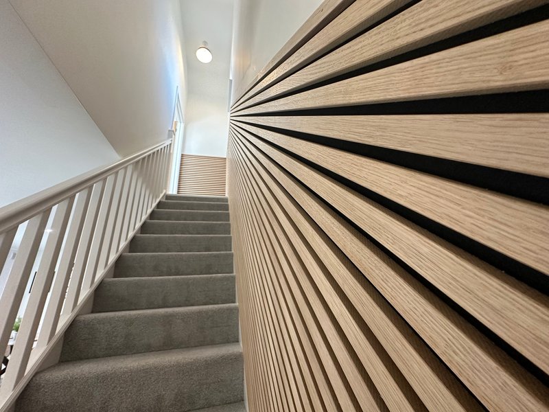 Modern staircase with wooden slatted wall paneling, metal handrail, and grey carpet treads