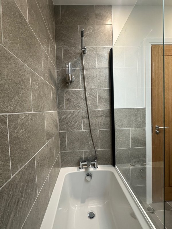 Modern bathroom with grey tile walls, white bathtub, glass shower screen and wooden door