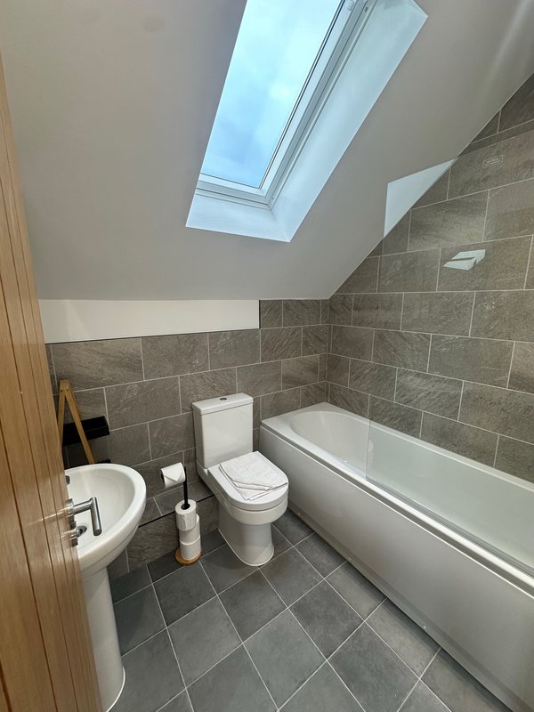 Modern bathroom with toilet, pedestal sink, bathtub, grey tile walls and skylight