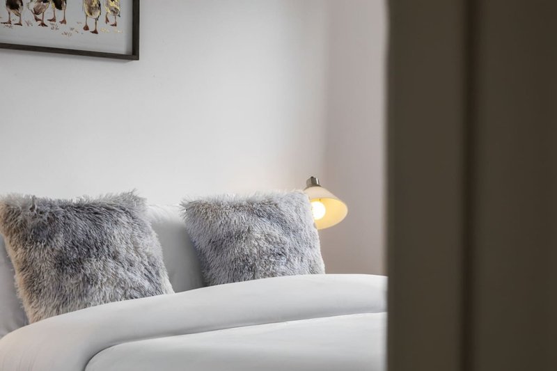 Modern bedroom with white bed, grey fluffy cushions and wall-mounted reading lamp