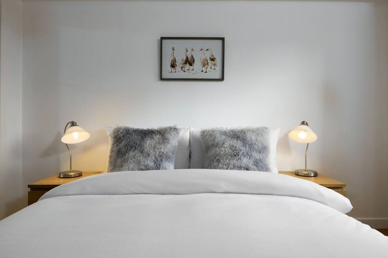 Bedroom with white duvet, grey cushions, wooden bedside tables, brass lamps, and framed wall art