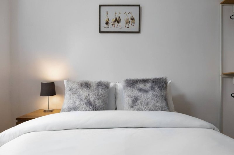 Bedroom with white bedding, gray fuzzy pillows, bedside lamp and framed wall art