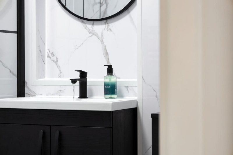 Modern bathroom vanity with black cabinetry, marble backsplash, round mirror and toiletries
