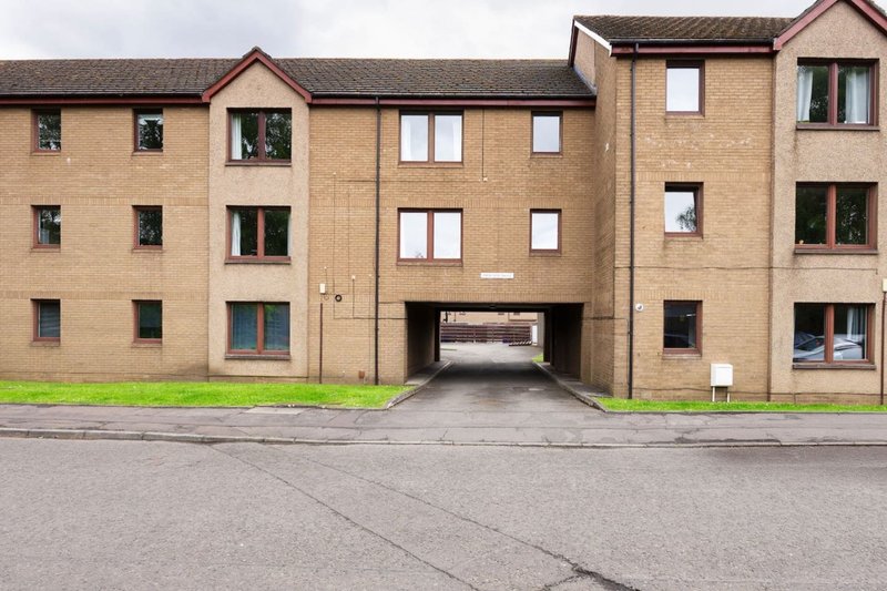 Brick apartment building exterior with multiple windows, central archway entrance and parking area