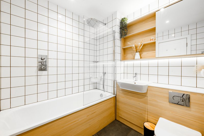 Modern bathroom with white tile walls, wooden vanity, bathtub, and shelving with decor