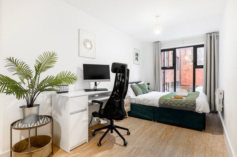 Modern bedroom with desk, office chair, bed, potted plant and large windows overlooking brick building