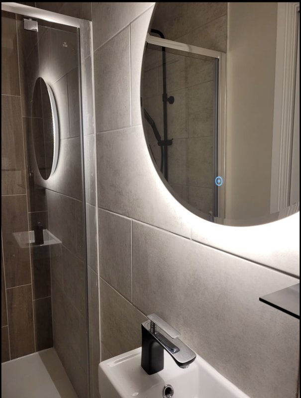 Modern bathroom with curved LED mirror, tiled walls, sink, and glass shower enclosure