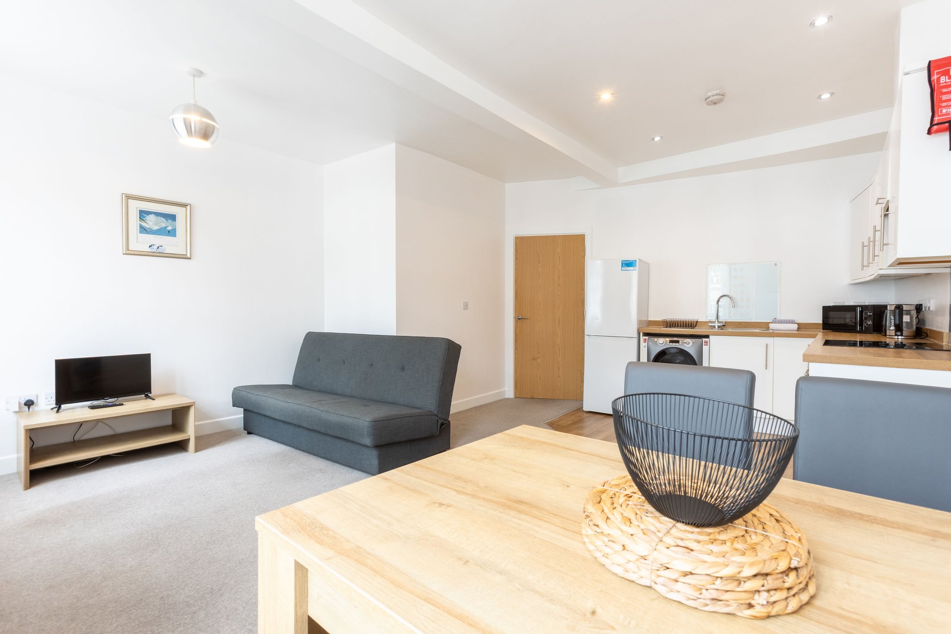 Open-plan living area with wooden dining table, grey sofa, TV, and kitchen units