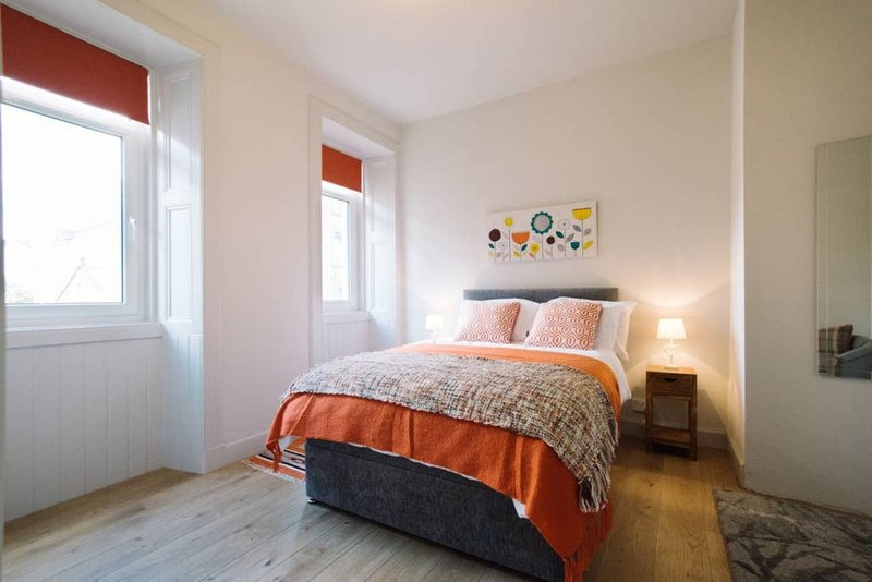 Bright bedroom with double bed, orange bedding, wooden floor and windows with red blinds