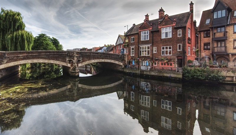 Historic brick buildings reflected in calm water with stone bridge overhead