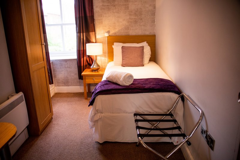 Compact bedroom with single bed, wooden headboard, bedside lamp and window with curtains
