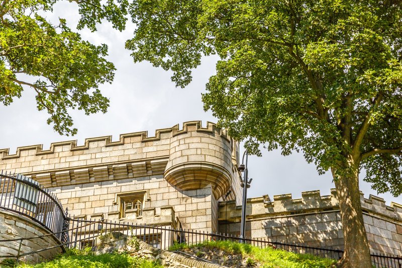 Stone castle exterior with crenellated walls, metal railings, and surrounding green trees