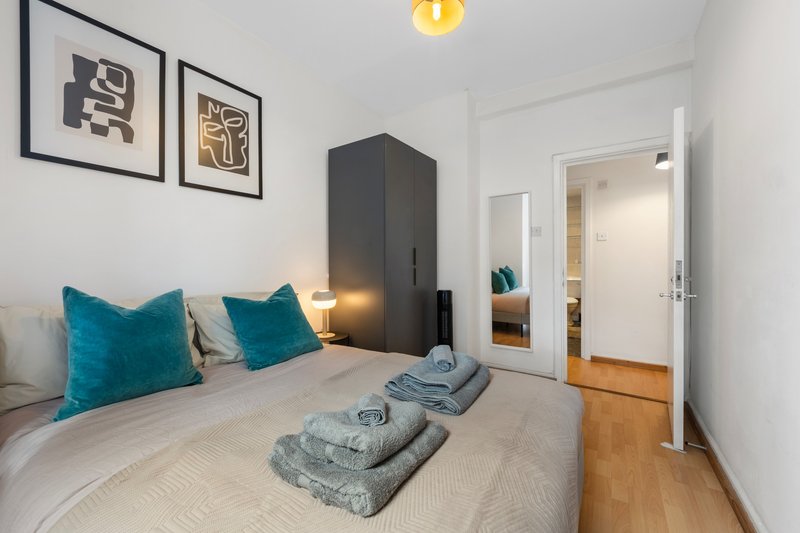 Modern bedroom with double bed, teal cushions, folded towels, dark wardrobe and framed wall art